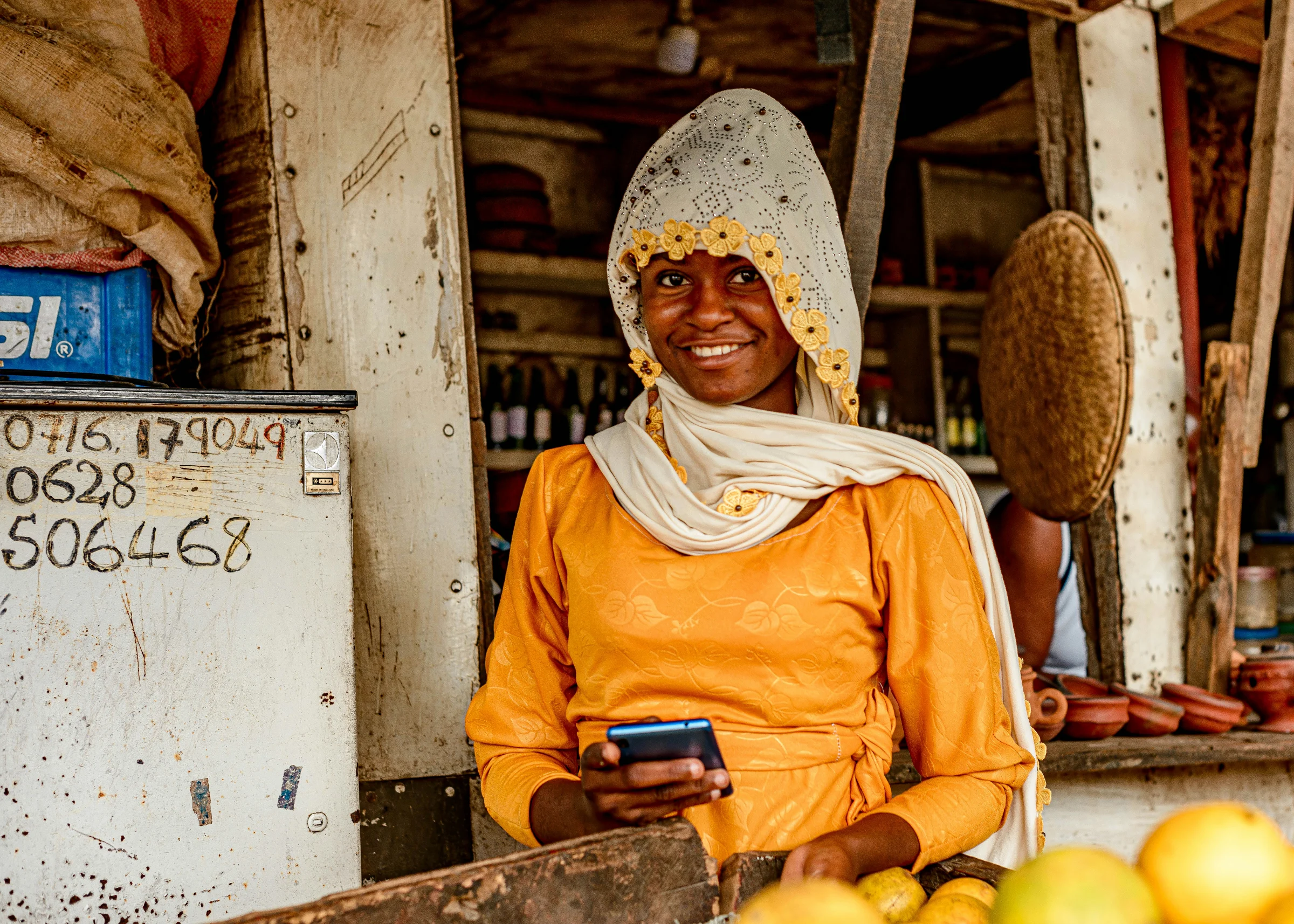 A woman uses a smartphone while working at a market stall, symbolizing the integration of modern technology into traditional livelihoods and daily commerce.